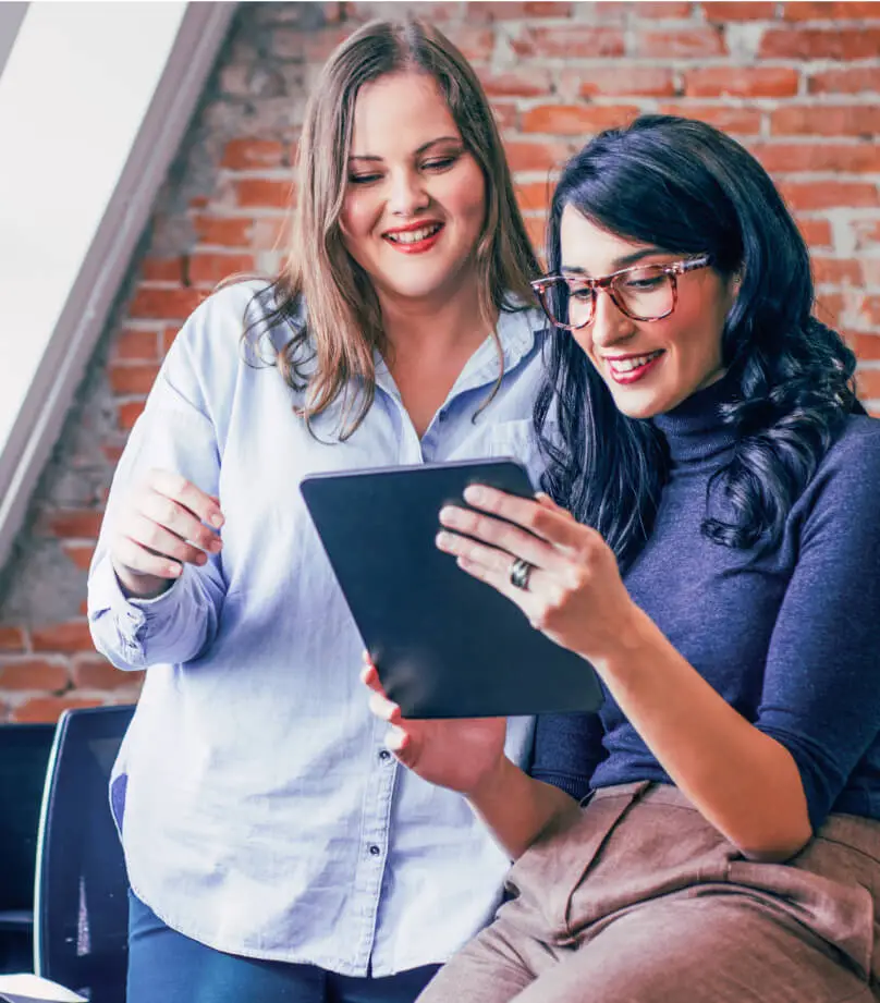 Two women smile while looking at a tablet.