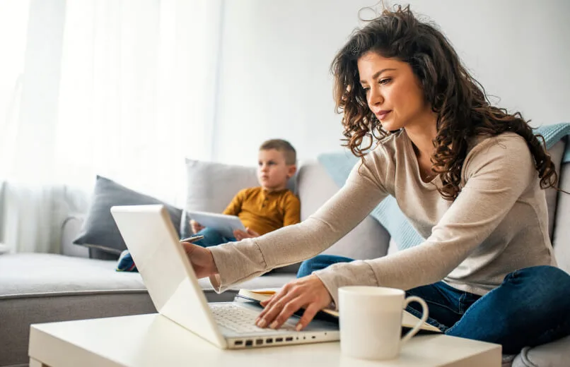 A woman sits on a couch looking at a laptop while a child sits on a couch in the distance with a tablet.