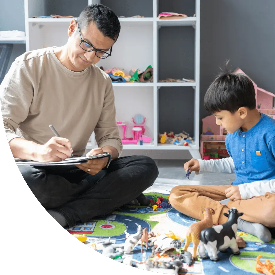 An adult with a notepad engages with a kid playing with minifigures on the ground.