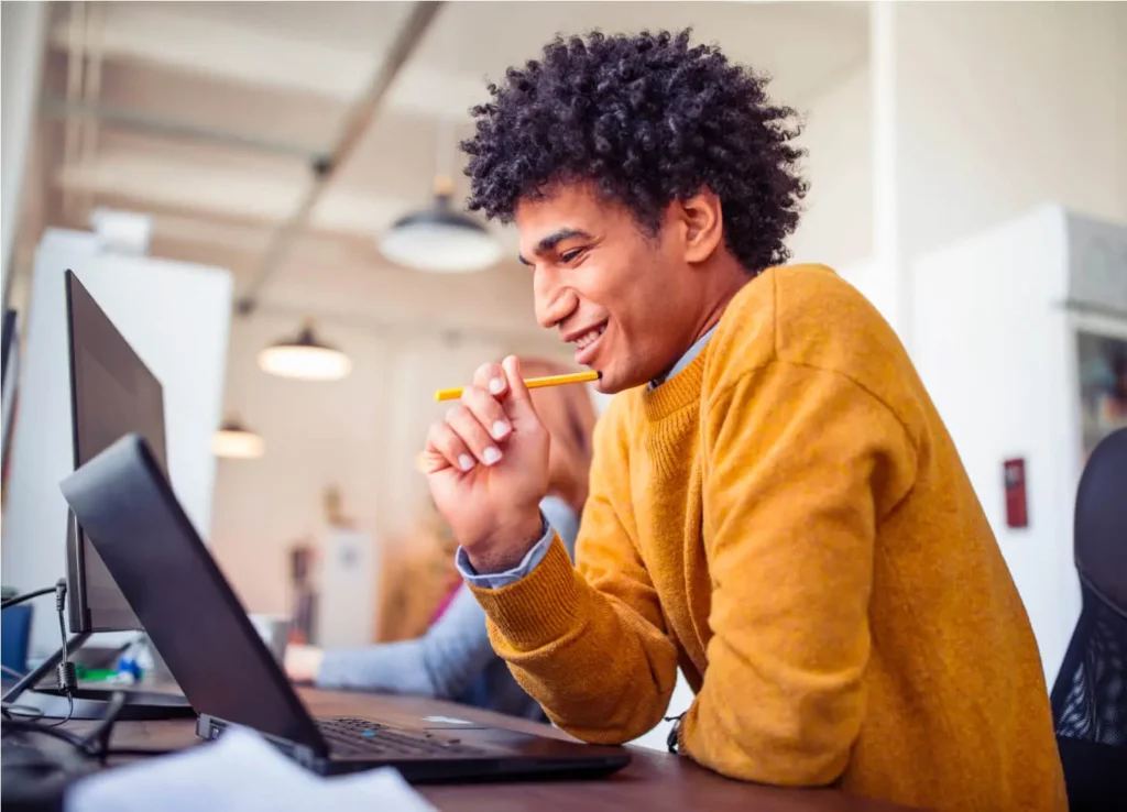 A man smile, holding a pencil against his chin while looking at a laptop screen.