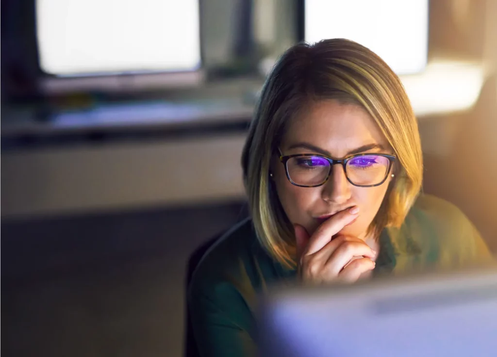A women in deep thought holds her hand over her mouth as she stars at the glow of the computer screen.