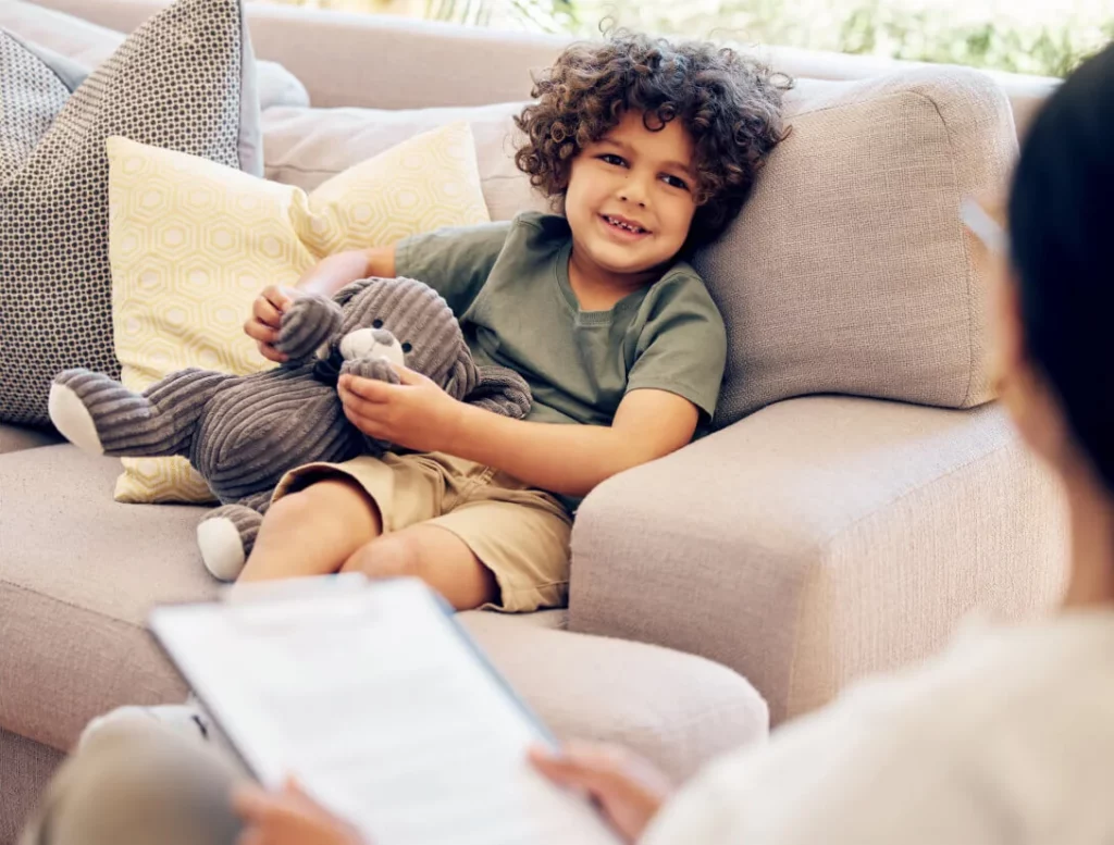 A child on a couch with a stuffed animal engages with an adult.