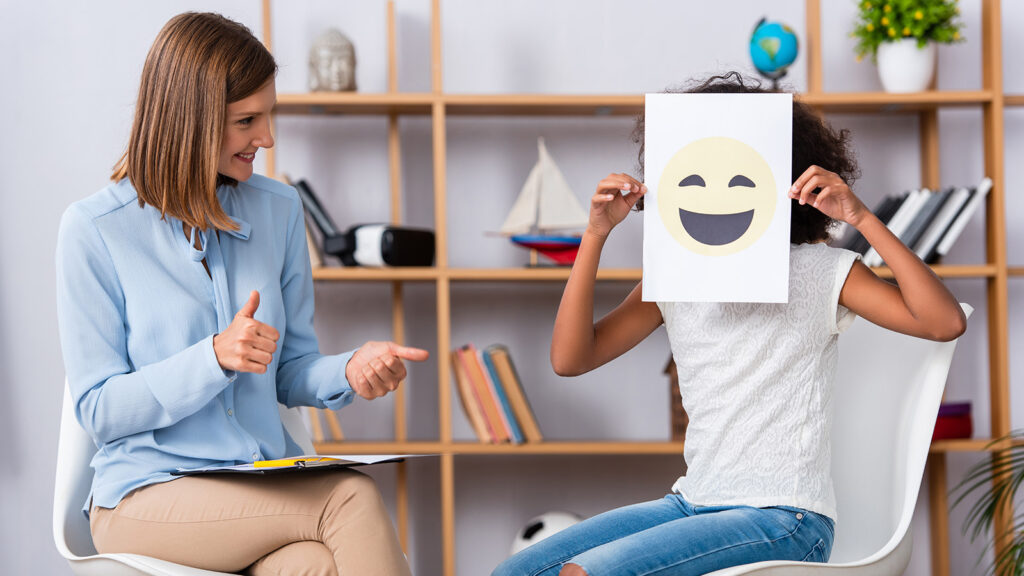 Smiling psychologist with thumbs up looking at african american girl covering face with happy expression on paper during consultation