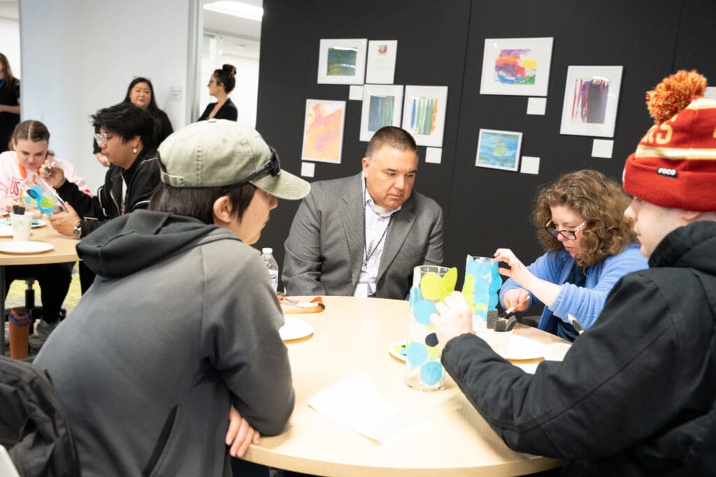 Multiple people sitting at tables while some work on crafts.  A man in a suit looks on.