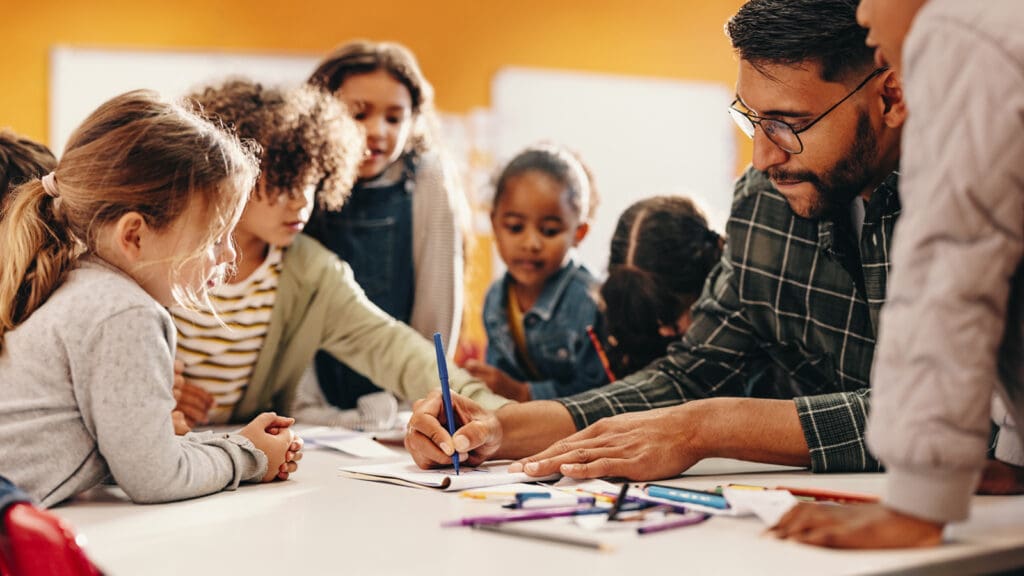 A diverse group of young children gather around a teacher at a table, intently watching him draw with a blue marker. Bright, warm classroom setting.